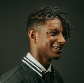 Smiling man with dreads in a varsity jacket.